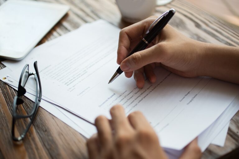 Unrecognizable businessman examining papers at table. Manager with ballpoint pen filling business papers. Close-up of male hands working at desk. Analyzing documents concept